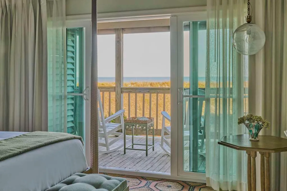 The image showcases the inviting interior of a studio with open doors leading to a private deck. On the deck, two classic rocking chairs face the Atlantic Ocean, inviting guests to relax and soak in panoramic water views. A decorative table with fresh flowers adds a touch of elegance and warmth to the serene, sunlit setting.