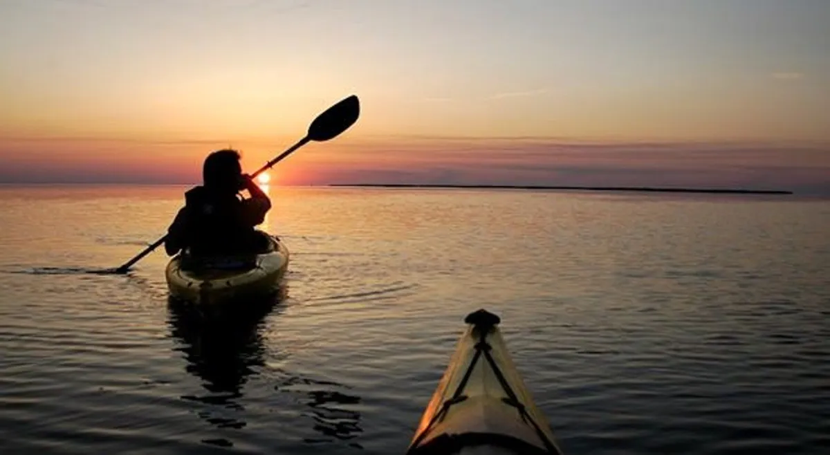 A kayaker glides across calm water at sunset, surrounded by a tranquil landscape. The golden light reflects off the water, enhancing the sense of adventure and natural beauty available just steps from the resort.