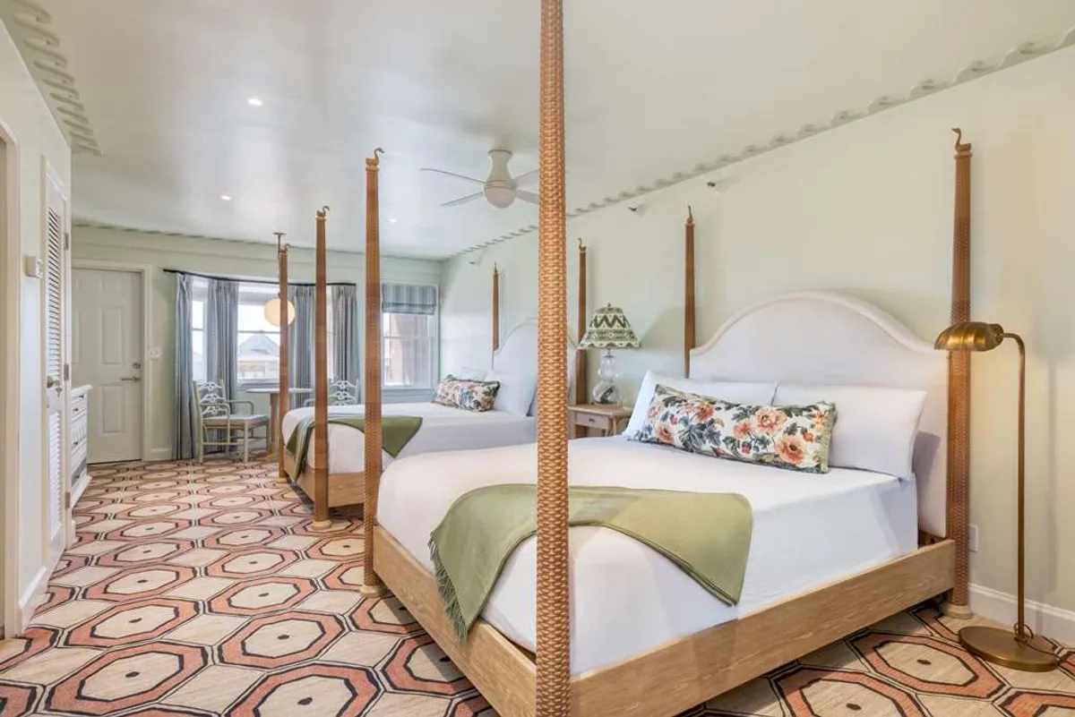 A bright, inviting guestroom featuring two four-poster beds topped with crisp linens, a decorative rug, and a colorful lamp. Natural light streams in, accentuating the comfortable and cheerful atmosphere—ideal for relaxation and restful sleep.