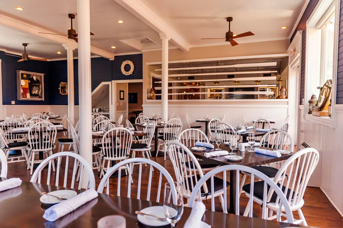 This image captures an interior dining venue with tables and white chairs arranged for guests, a visible bar area in the background, and decorative wall art. The atmosphere is casual yet lively, with people engaging in dining and socializing, signaling a comfortable environment that welcomes families and groups for memorable meals.
