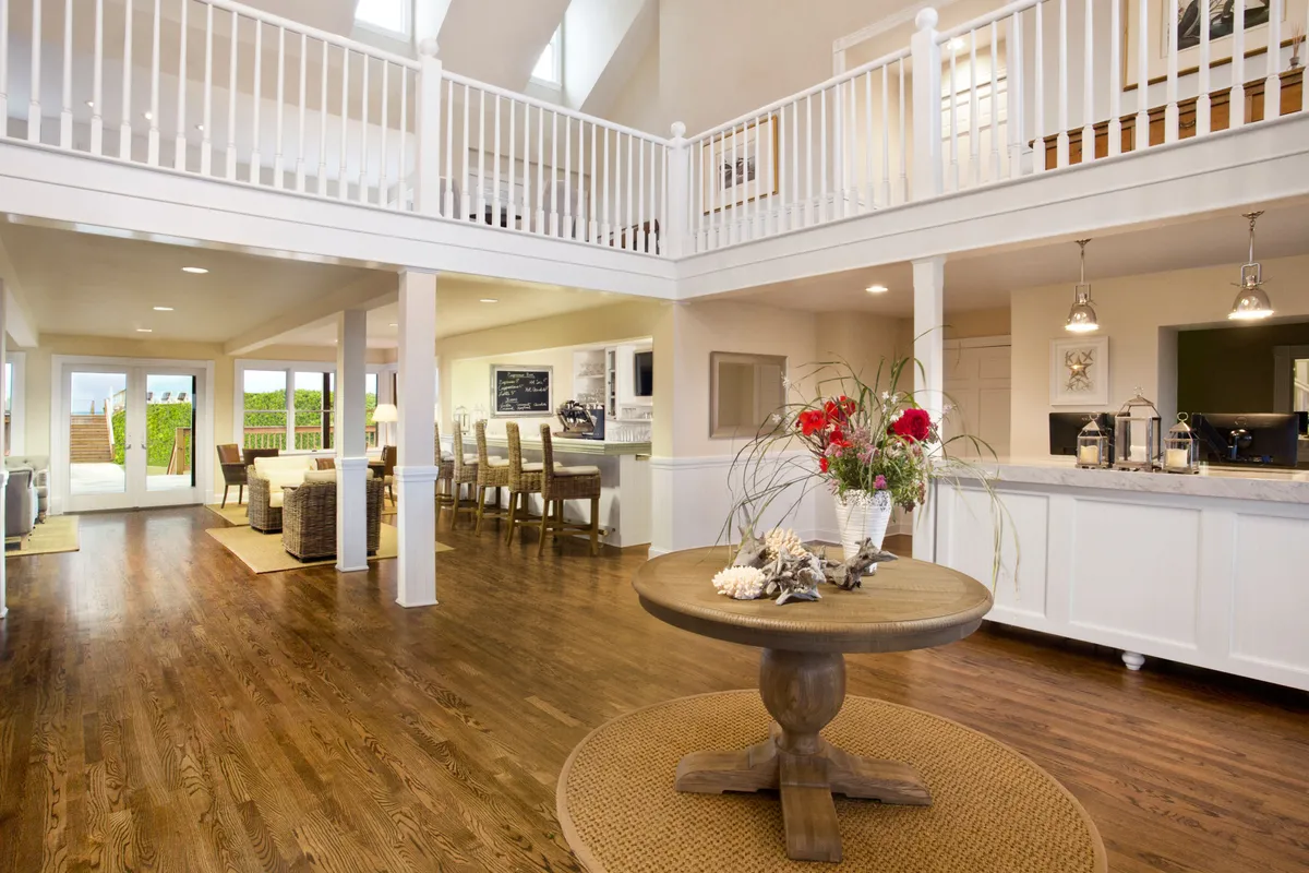 A bright, homey interior with a central round table adorned with a floral arrangement. The open floor plan leads to a dining area and kitchen, with large windows framing outdoor views. The setting feels perfect for board games, reading, and indoor gatherings on a rainy day.