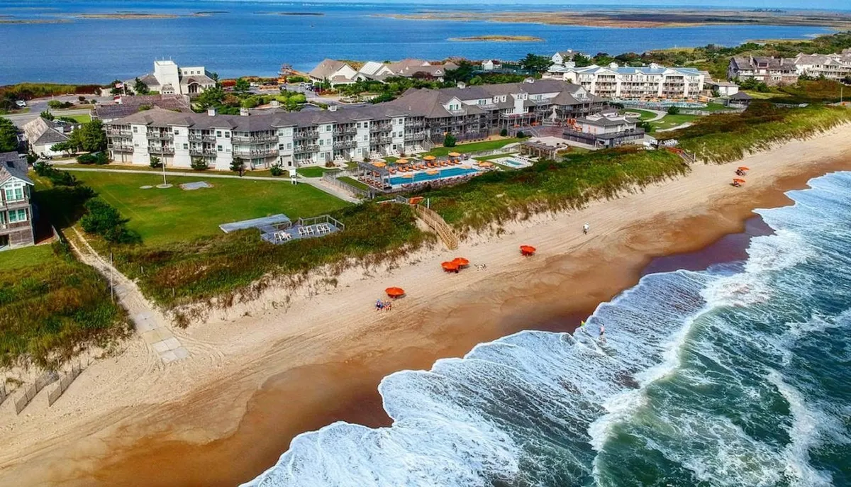 A wide-angle perspective displays the beachfront hotel, a glistening swimming pool, and rows of umbrellas on the sand—seamlessly blending the pool area, hotel amenities, and direct beach access. The scene is animated by guests swimming, sunbathing, and gathering by the water's edge.