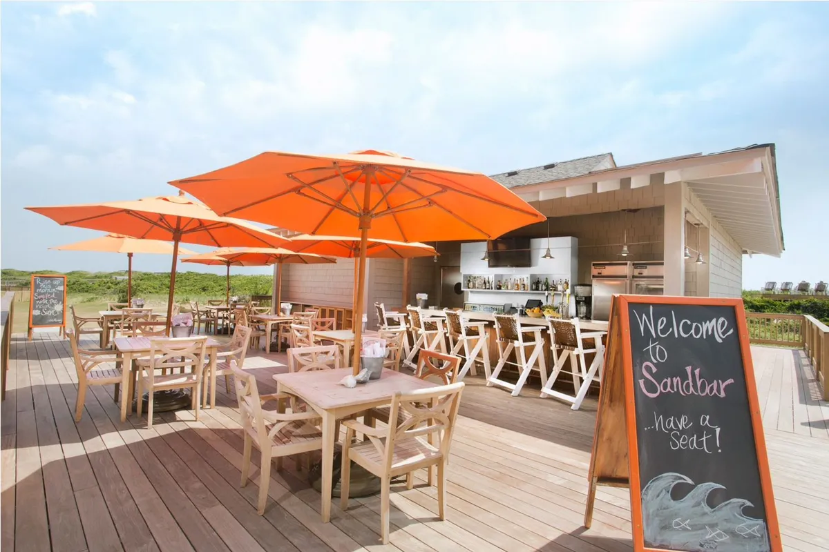 An outdoor dining area with orange umbrellas and wooden tables, set along the beach with a lively, relaxed atmosphere. The bar counter and sunset lighting set the stage for enjoying drinks or a casual meal, just steps from the water.