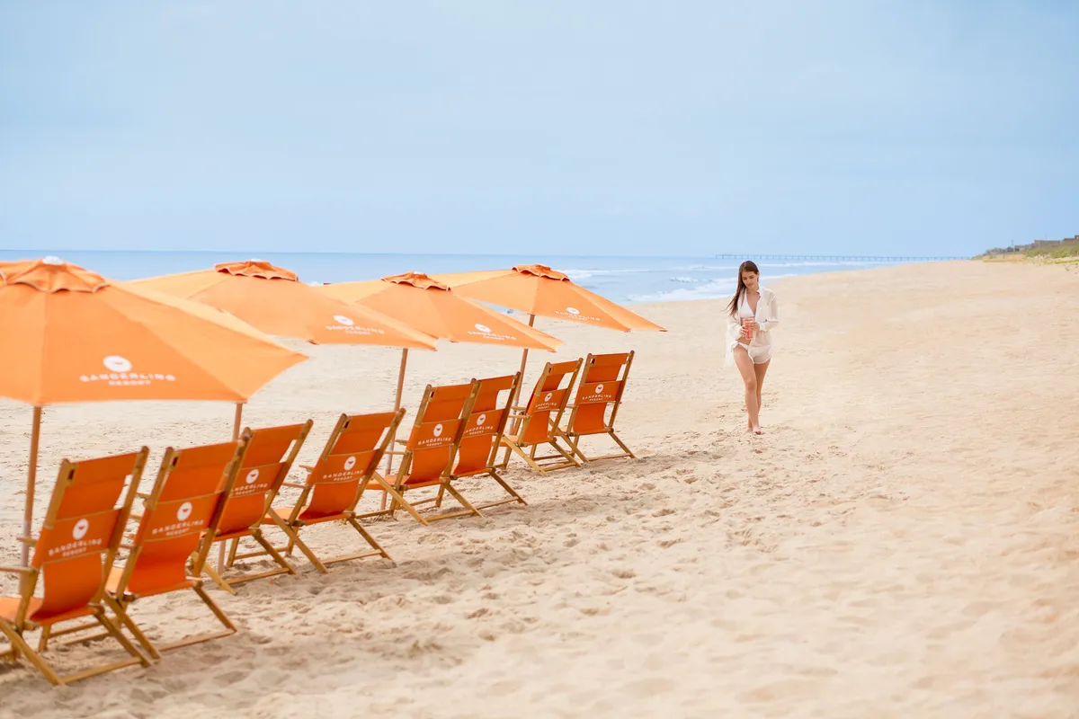 A serene stretch of private beach with neatly arranged chairs and umbrellas facing the ocean. A person strolls along the shore, enjoying the peaceful ambiance, while the gentle waves and golden sunset hint at a relaxed, communal resort experience.