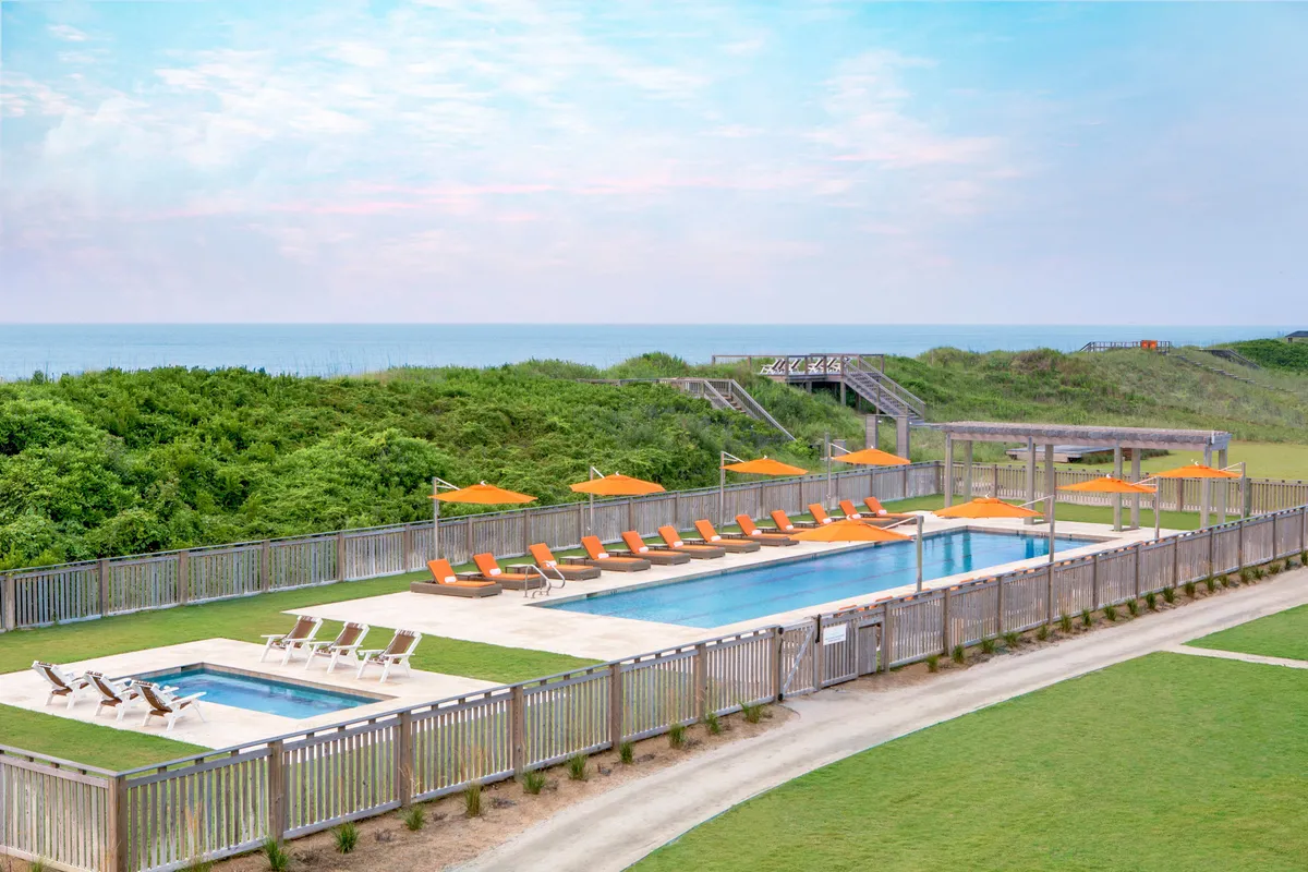 This vibrant exterior image showcases a spacious swimming pool surrounded by lounge chairs and orange umbrellas, set against lush landscape greenery. Guests can be seen swimming, sunbathing, and gathering in small social groups. The tranquil ambiance is enhanced by the soft glow of a sunset reflecting off the water.