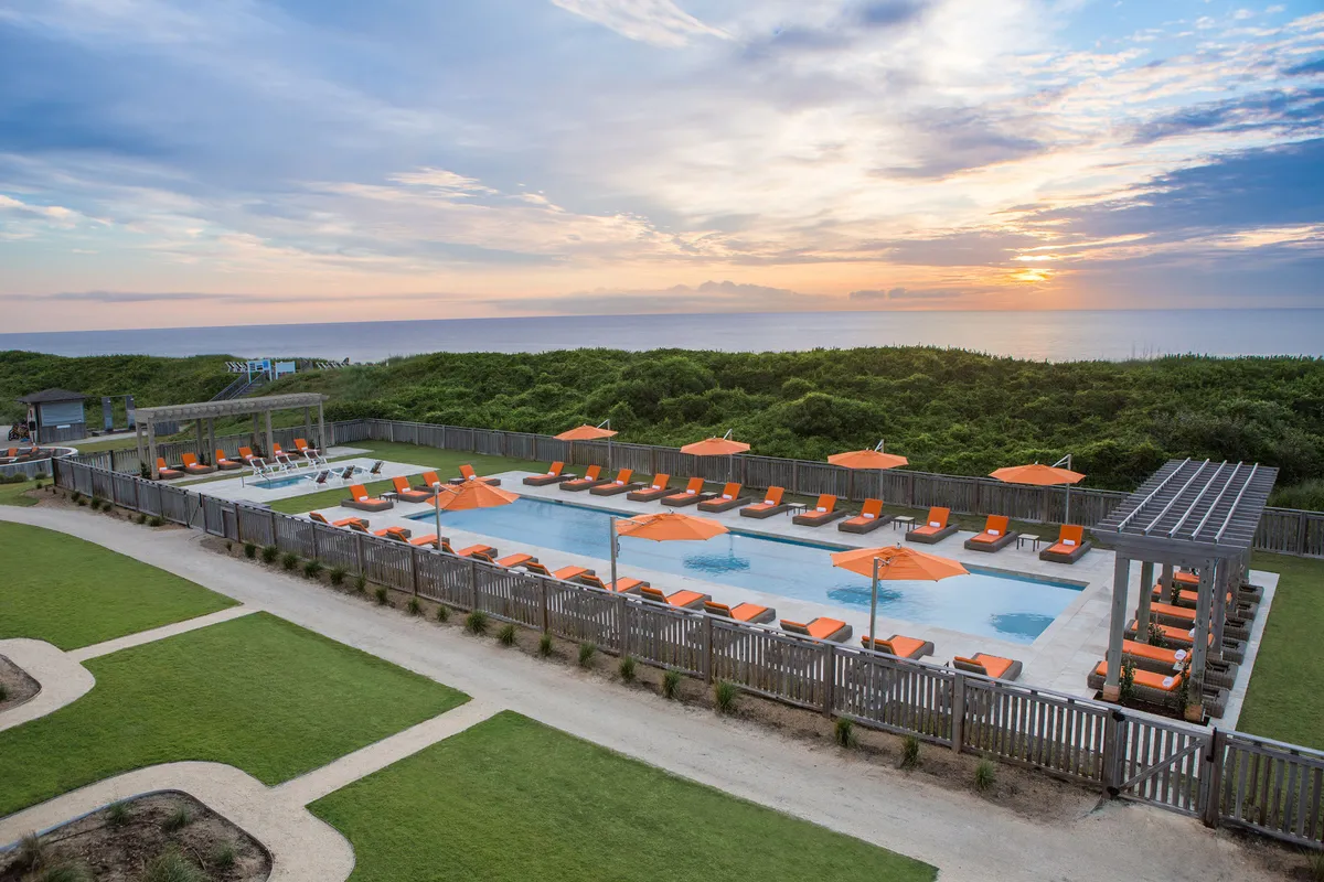A sun-drenched pool area overlooking the ocean, with sun loungers and orange umbrellas arranged invitingly. The tranquil mood is enhanced by breathtaking water views, ideal for post-workout relaxation or a leisurely sunset swim.