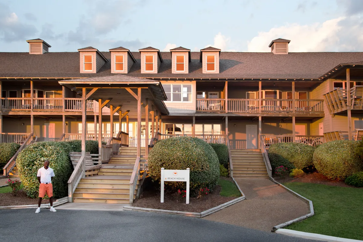 A welcoming exterior shot of the beach house, featuring a charming porch with hammock chairs, manicured bushes, and steps leading up to the entrance. The golden light suggests a sunset moment, and the inviting porch is perfect for relaxing and taking in panoramic ocean views.