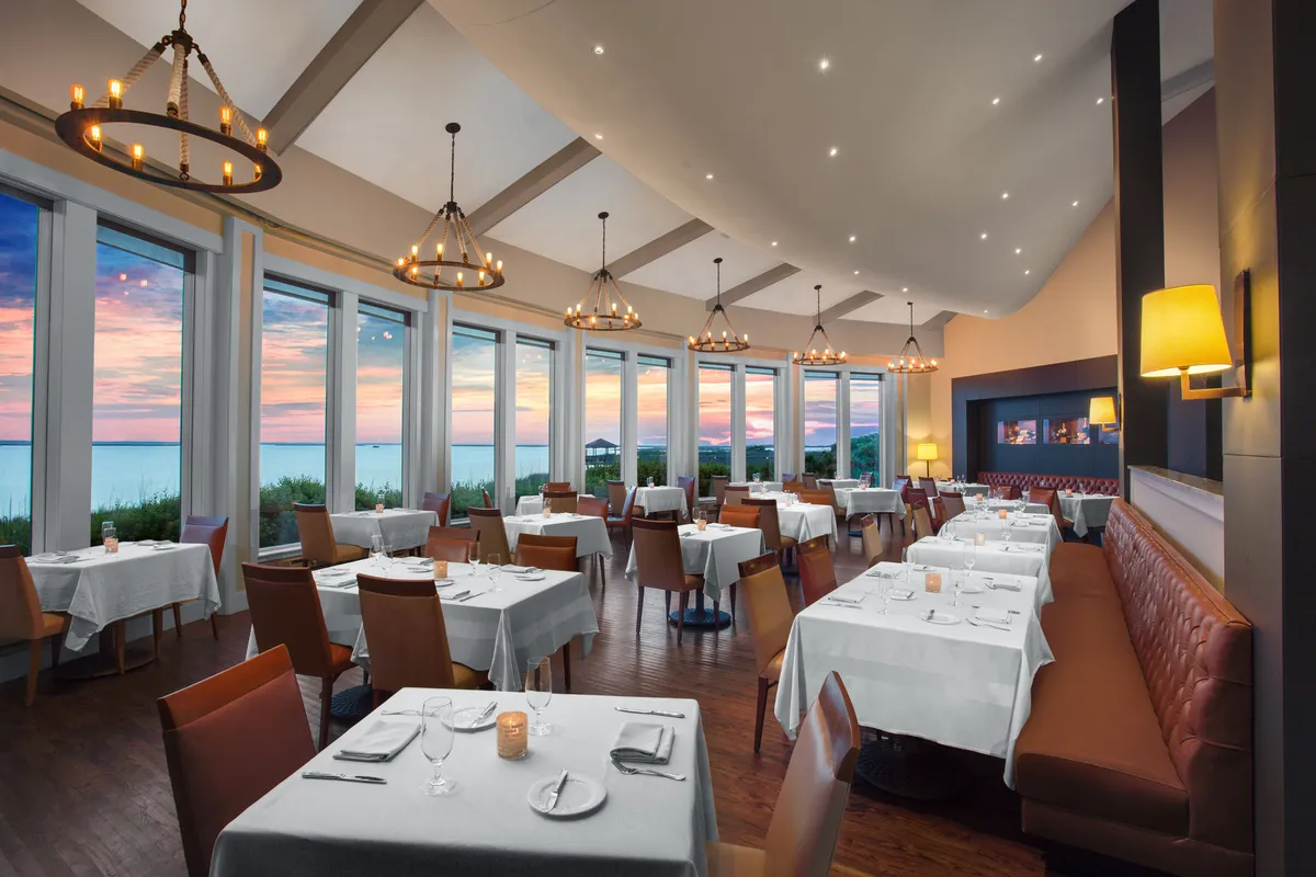 This image showcases a sophisticated dining space with large windows that offer a sunset view over the water. The tables are elegantly set with white linens, and the ambiance is elevated by beautiful chandeliers. The setting is perfect for refined dining, social gatherings, or romantic evenings, allowing guests to enjoy picturesque scenery while savoring healthy dishes.