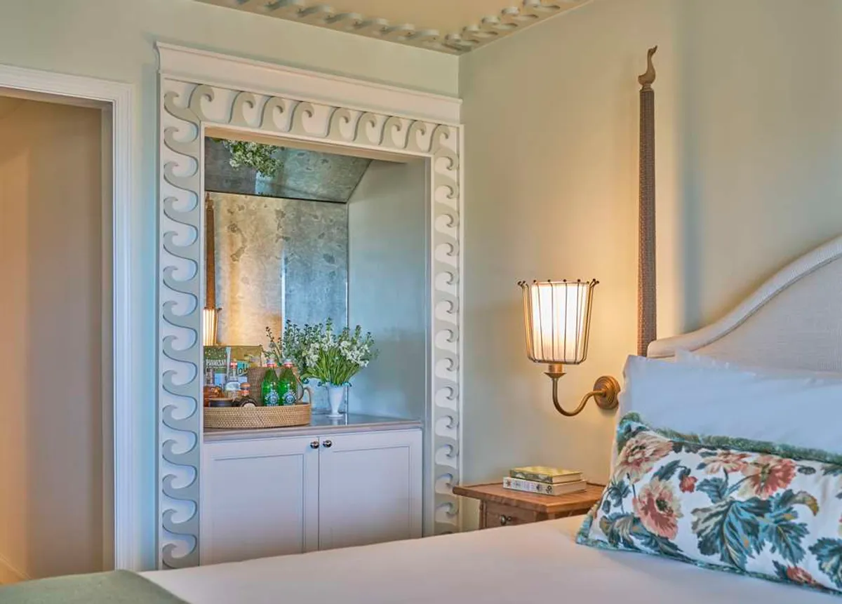 A serene guestroom vignette showing a bed with floral accents, a decorative mirror with an intricate frame, a vase of flowers, and a table lamp by a sunlit window. The atmosphere is tranquil, perfect for quiet moments with a book or coffee.
