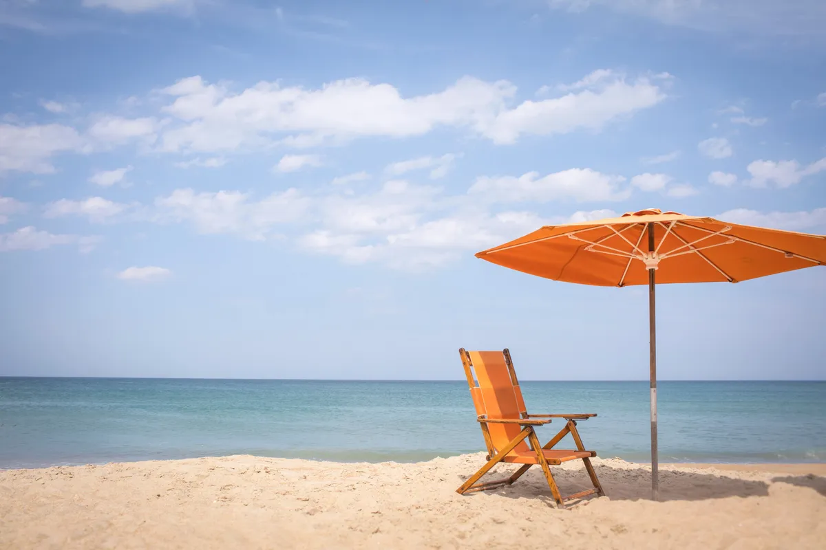 A calm, serene stretch of sandy beach with an orange beach chair and umbrella facing the tranquil ocean. The scene evokes sunny days, peaceful relaxation, and the potential for both solitary and family time by the water.