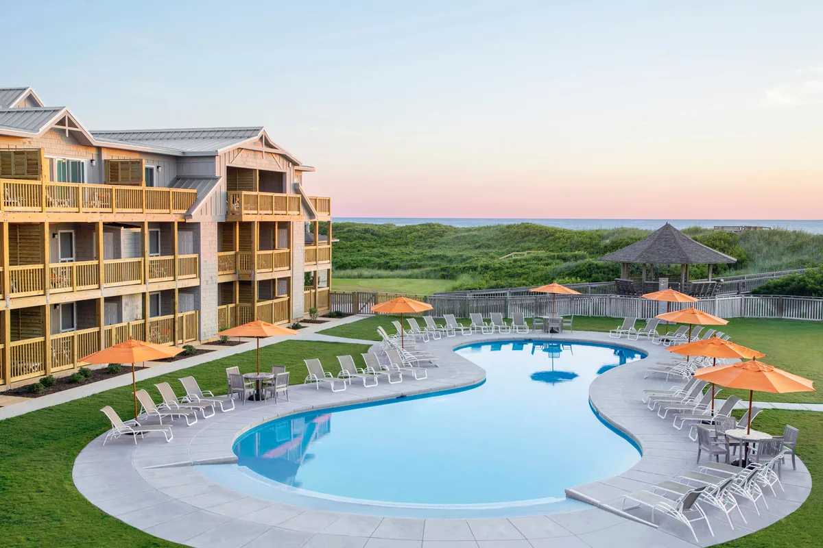 This image showcases a vibrant outdoor pool area with sparkling blue water, surrounded by orange umbrellas and a landscaped grass area. Balconies from the adjacent resort overlook the pool, and the inviting atmosphere hints at family fun, social gatherings, and relaxation beneath the evening sky.